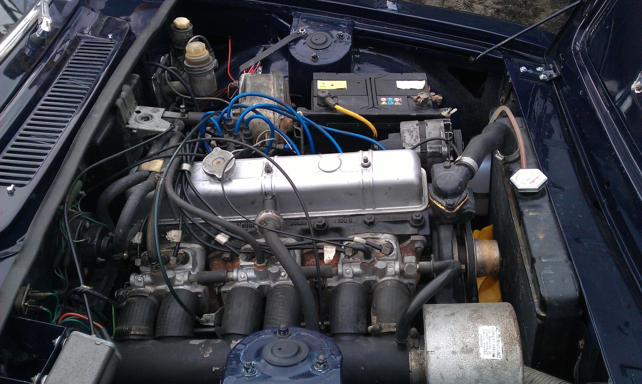 The engine bay of a dark blue saloon: a six-cylinder engine with six short intake pipes and injection lines along its side.