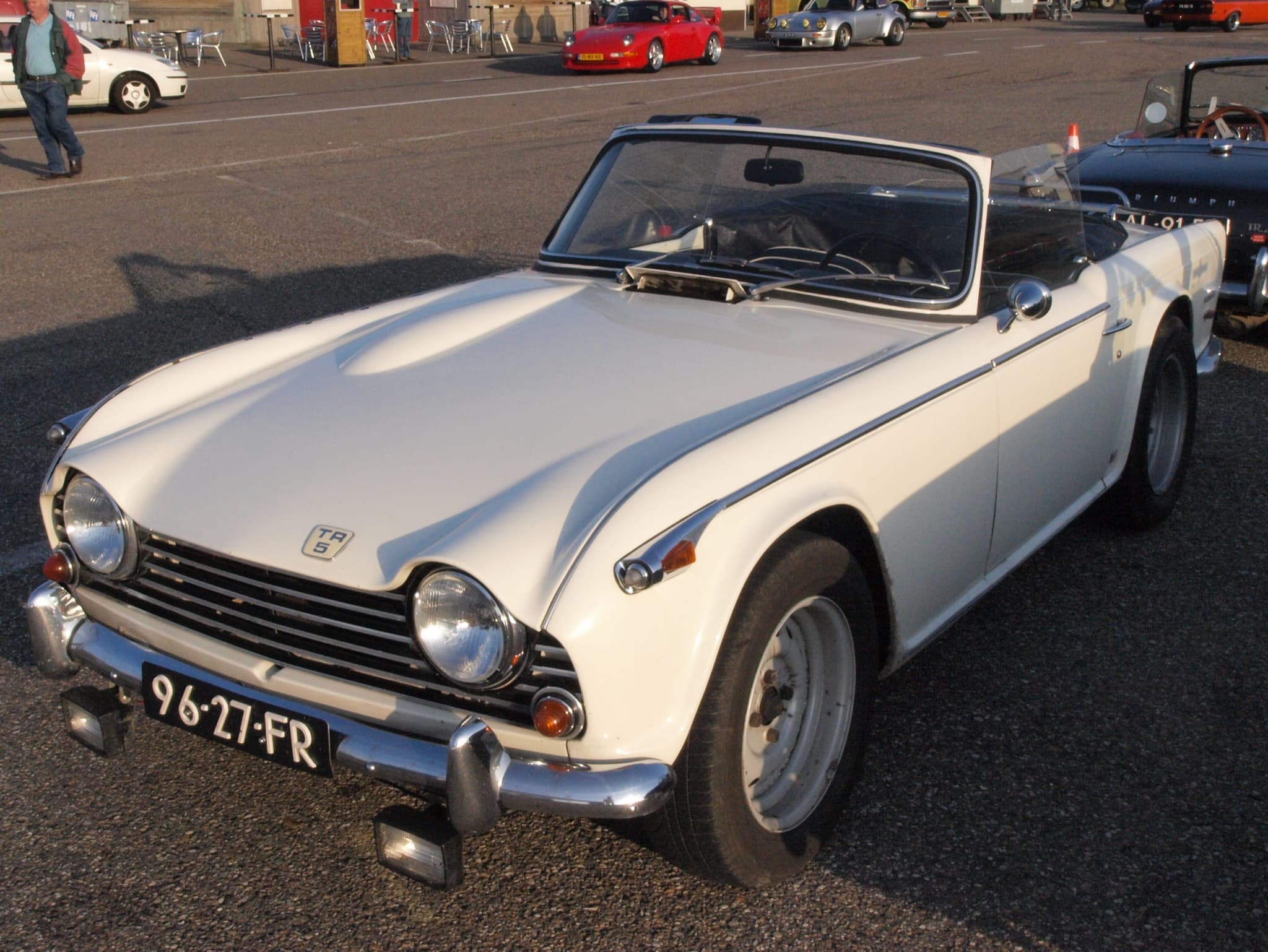 A white sports car with its hood folded and square lamps under the bumper, on tarmac in low evening sun. Opens a larger view
