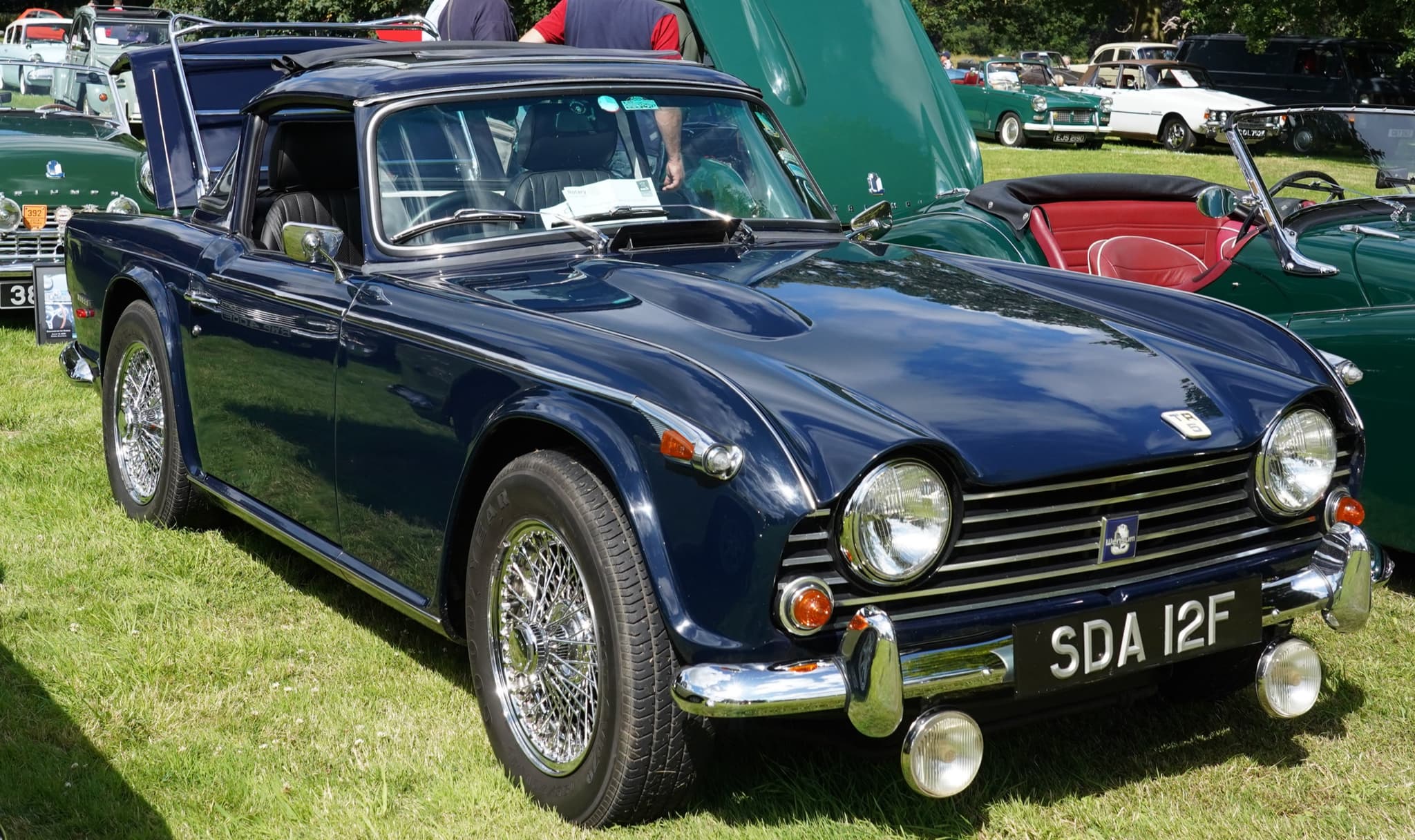 A dark blue sports car with a hardtop frame, a black fabric roof panel, wire wheels and spot lamps, on grass at a show. Opens a larger view