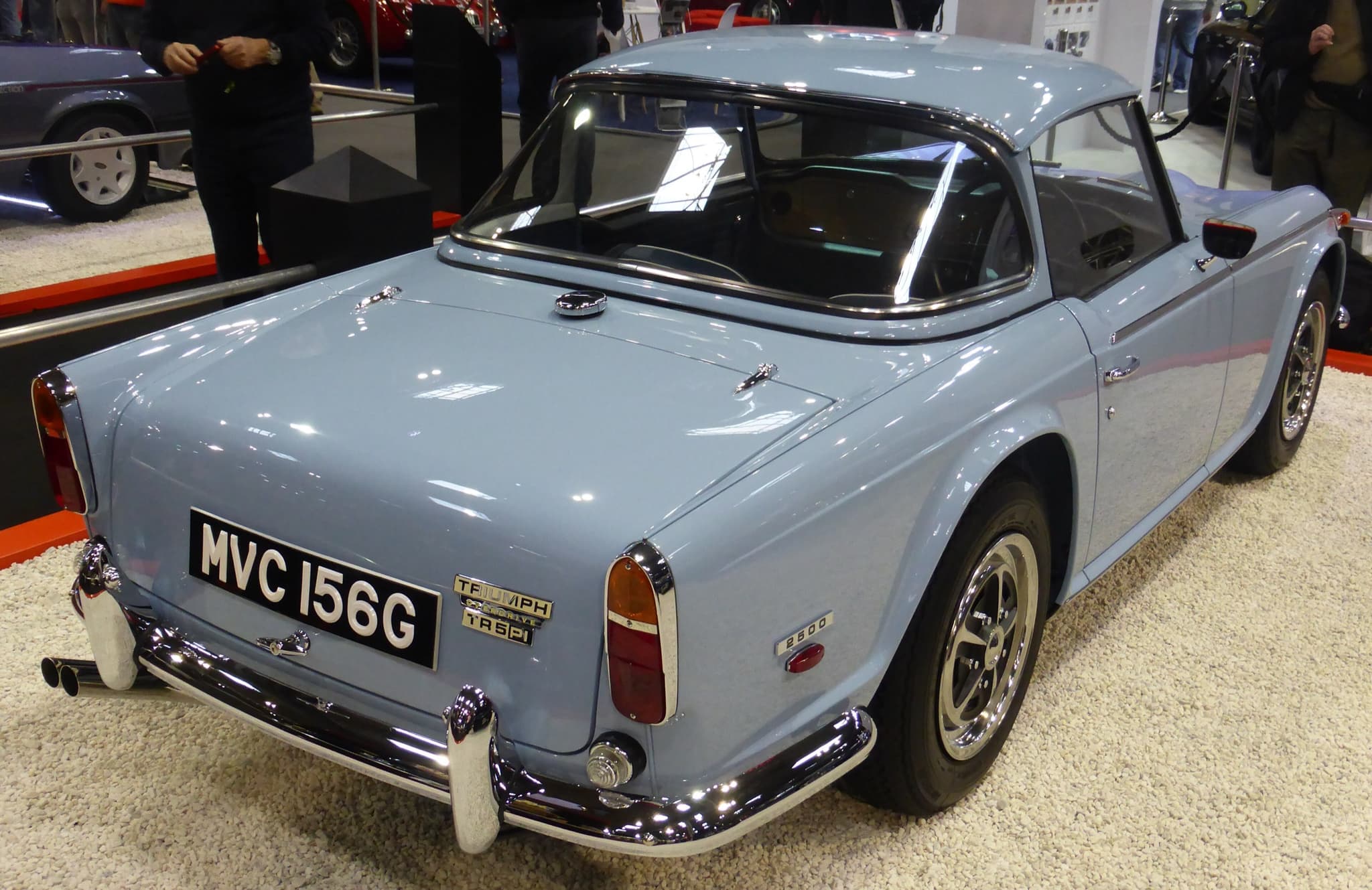 A pale blue sports car with a matching hardtop seen from behind on a show stand, its badges and twin exhausts in view. Opens a larger view