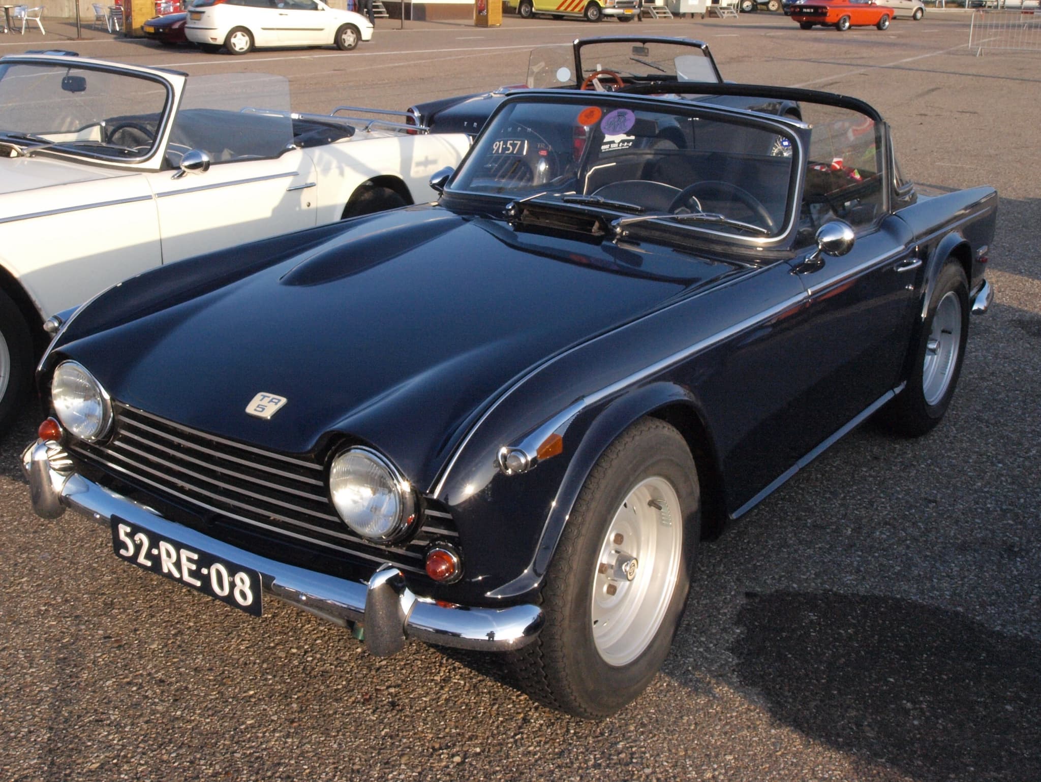A dark blue sports car with its hood folded, front three-quarter on tarmac in low evening sun beside a white roadster. Opens a larger view