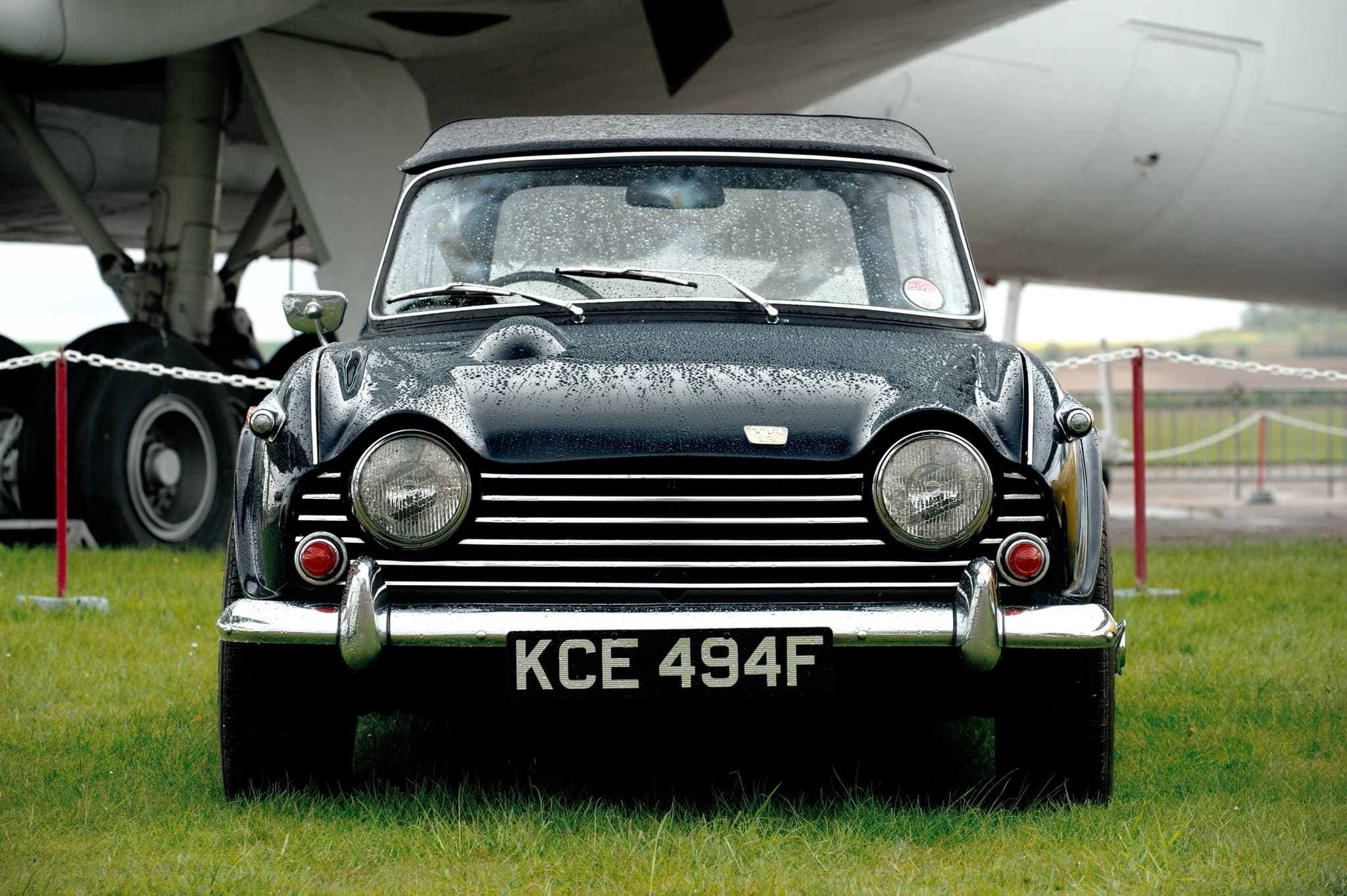 A black sports car with its hood up, beaded with rain, parked head-on on grass beneath the wing of a large airliner. Opens a larger view