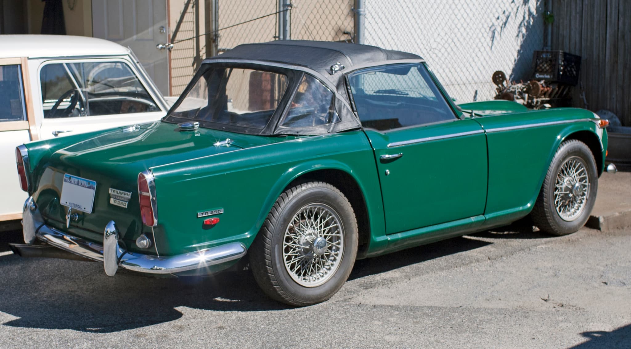 A green open sports car with its black hood up, seen from behind, with TR250 badges on the boot and rear wing.