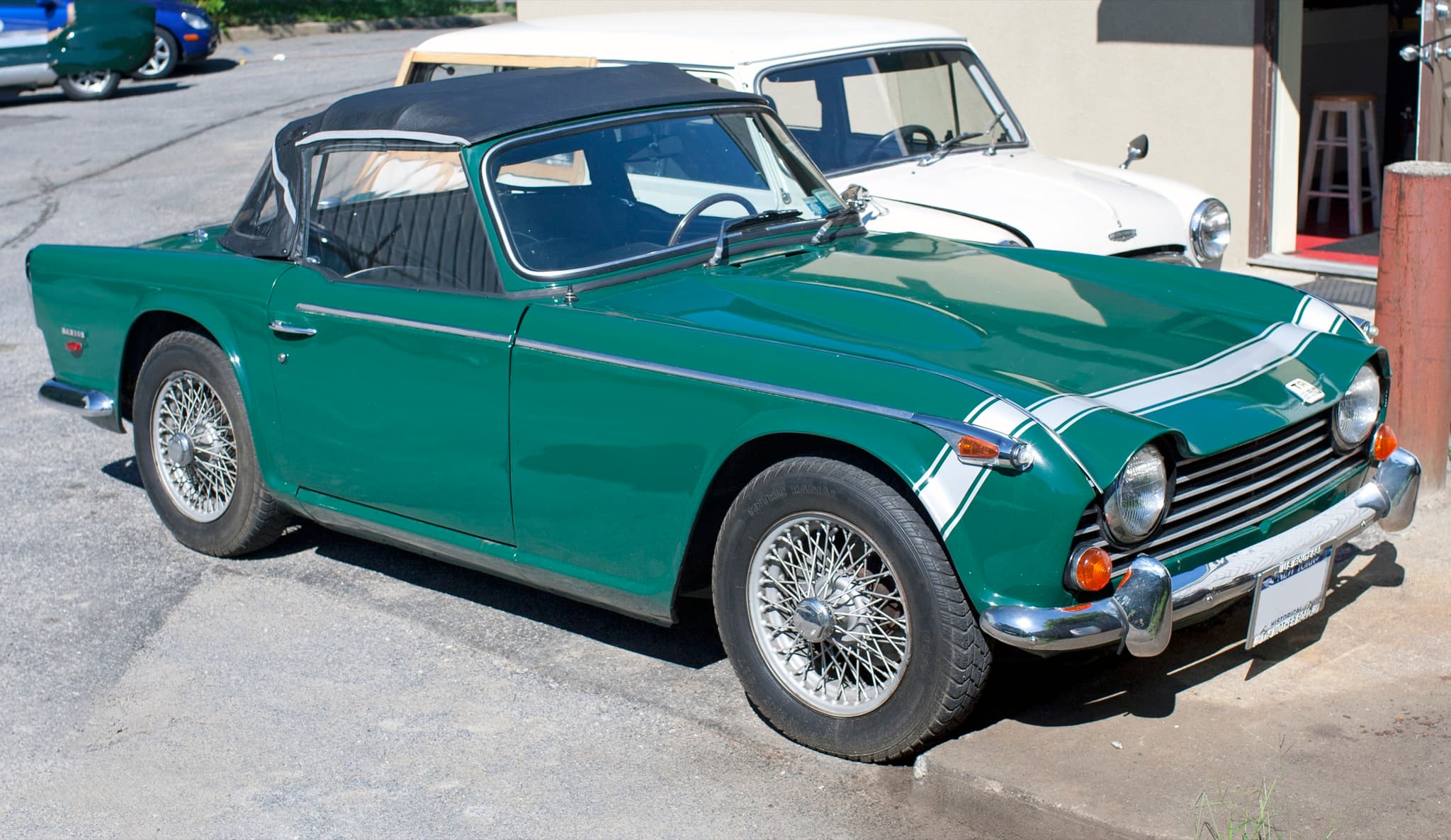 A green Triumph TR250 with a white stripe across its nose and its black hood up, seen from the front three-quarter in a sunny yard.