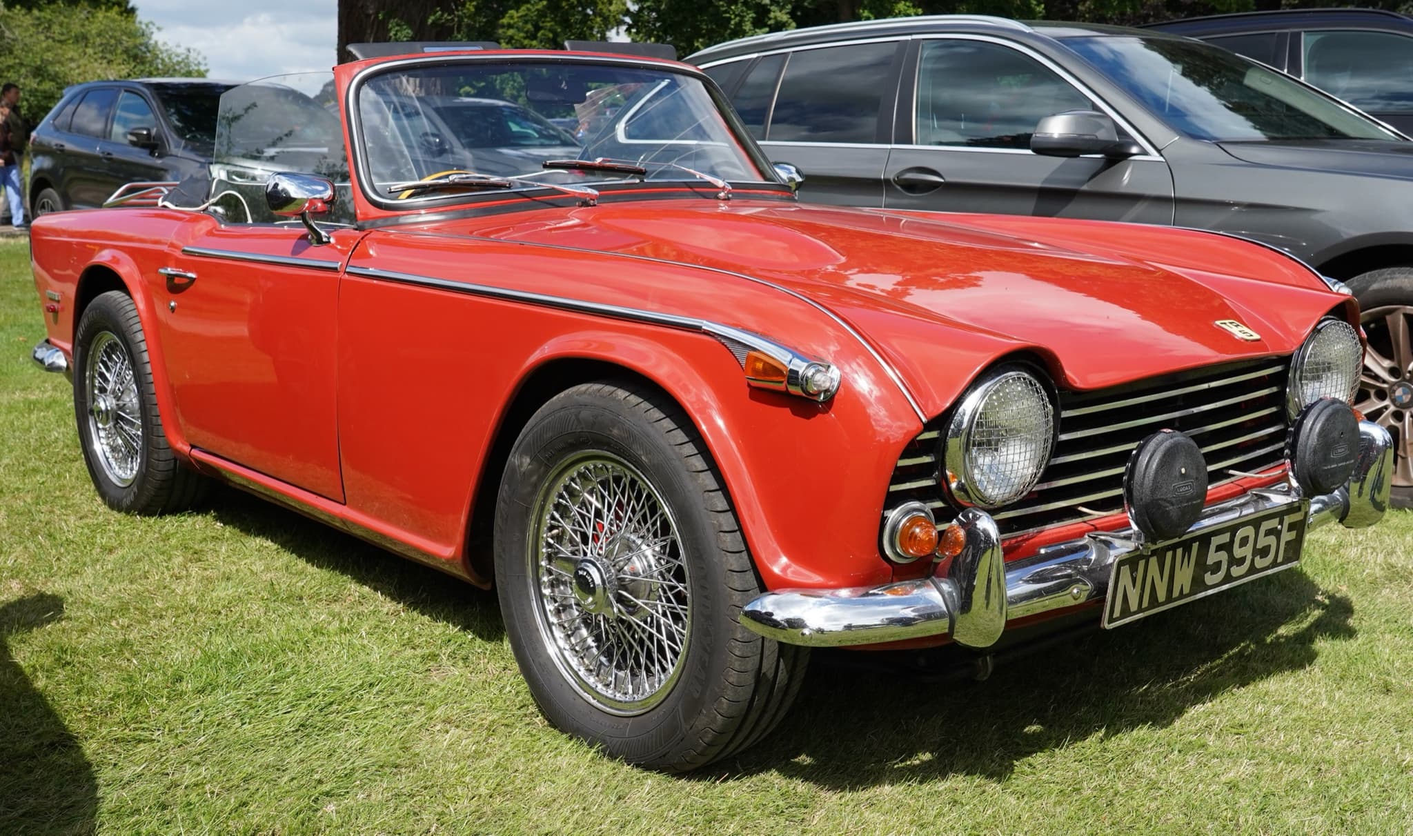 A red sports car with wire wheels and two spot lamps on its bumper, parked on grass in front of modern cars.