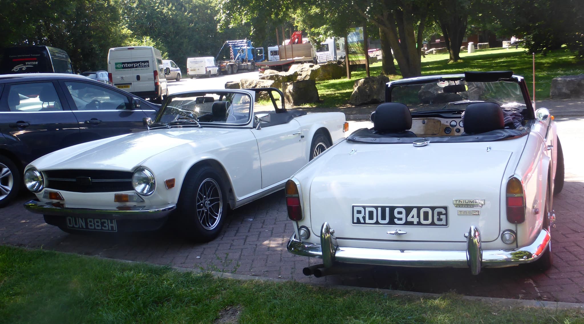 A white sports car seen from behind parked nose to nose with a white sports car with a squarer front, on block paving.