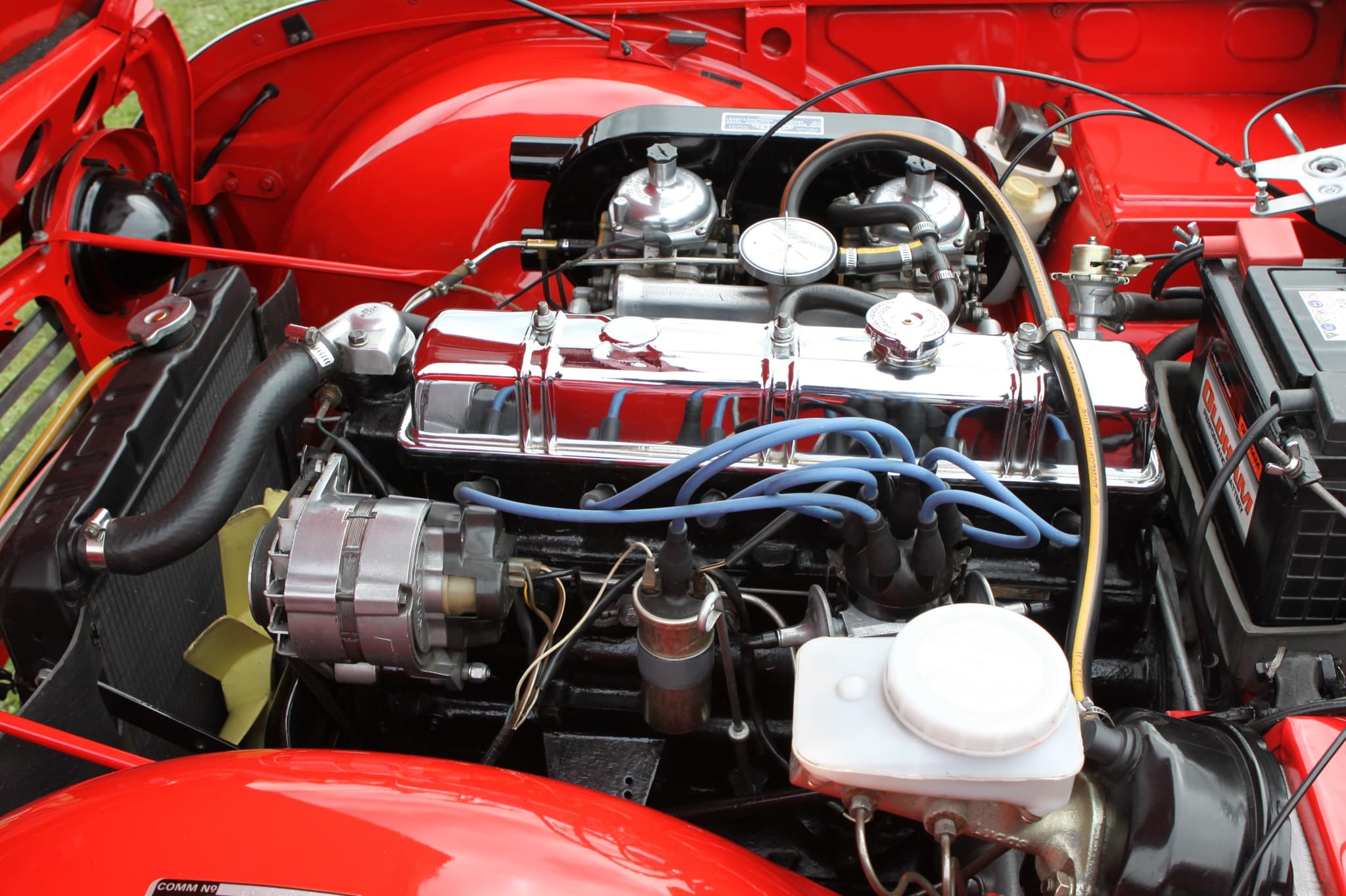 The engine bay of a red car: a six-cylinder engine with a chromed rocker cover, blue plug leads and two carburettors behind.