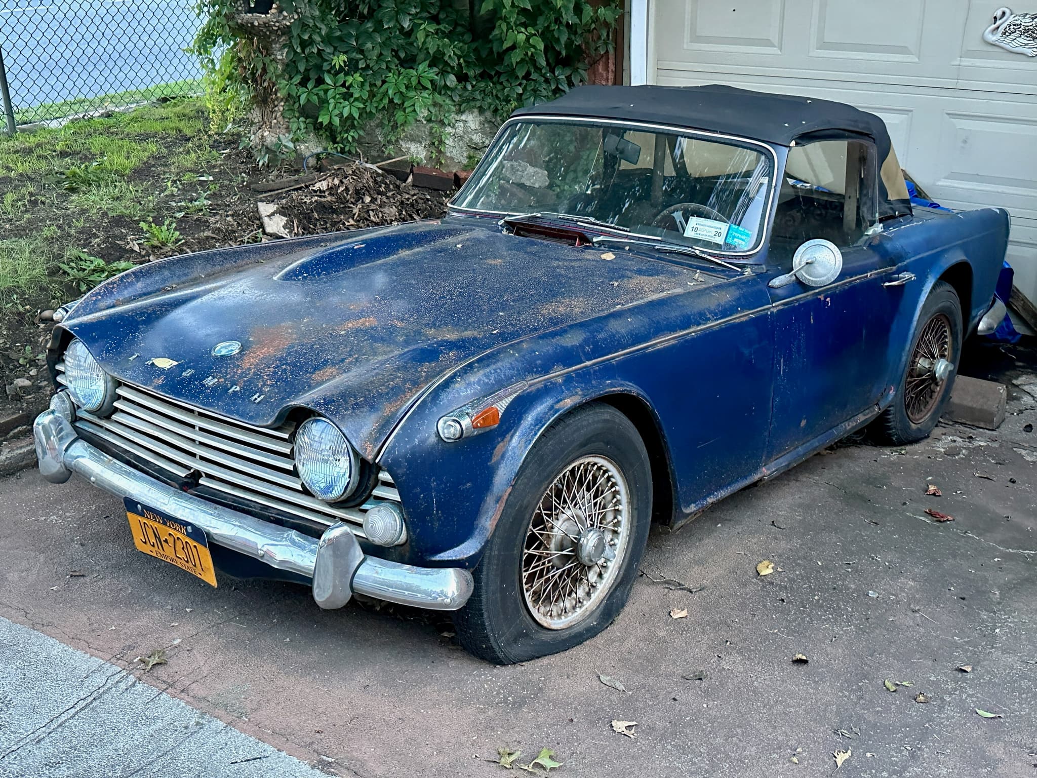 A neglected dark blue sports car with faded, rusting paint, flat tyres and leaves on the bonnet, parked by a garage door.