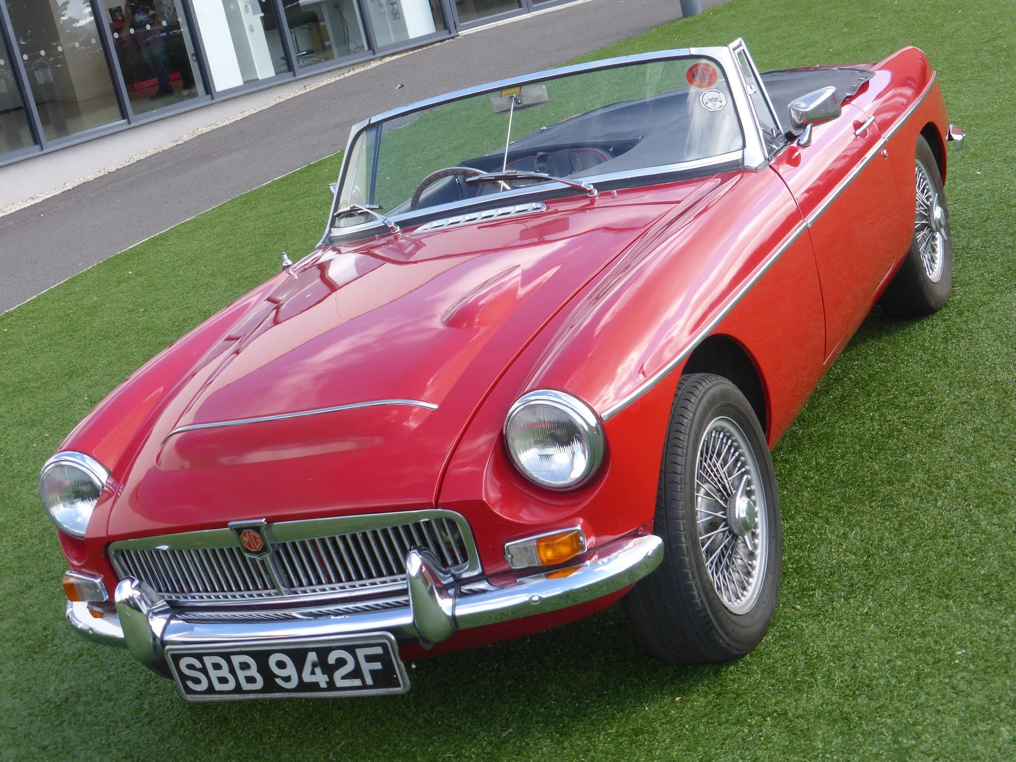 A red MGC roadster with wire wheels and a bulged bonnet, parked on artificial grass in front of a glass building. Opens a larger view