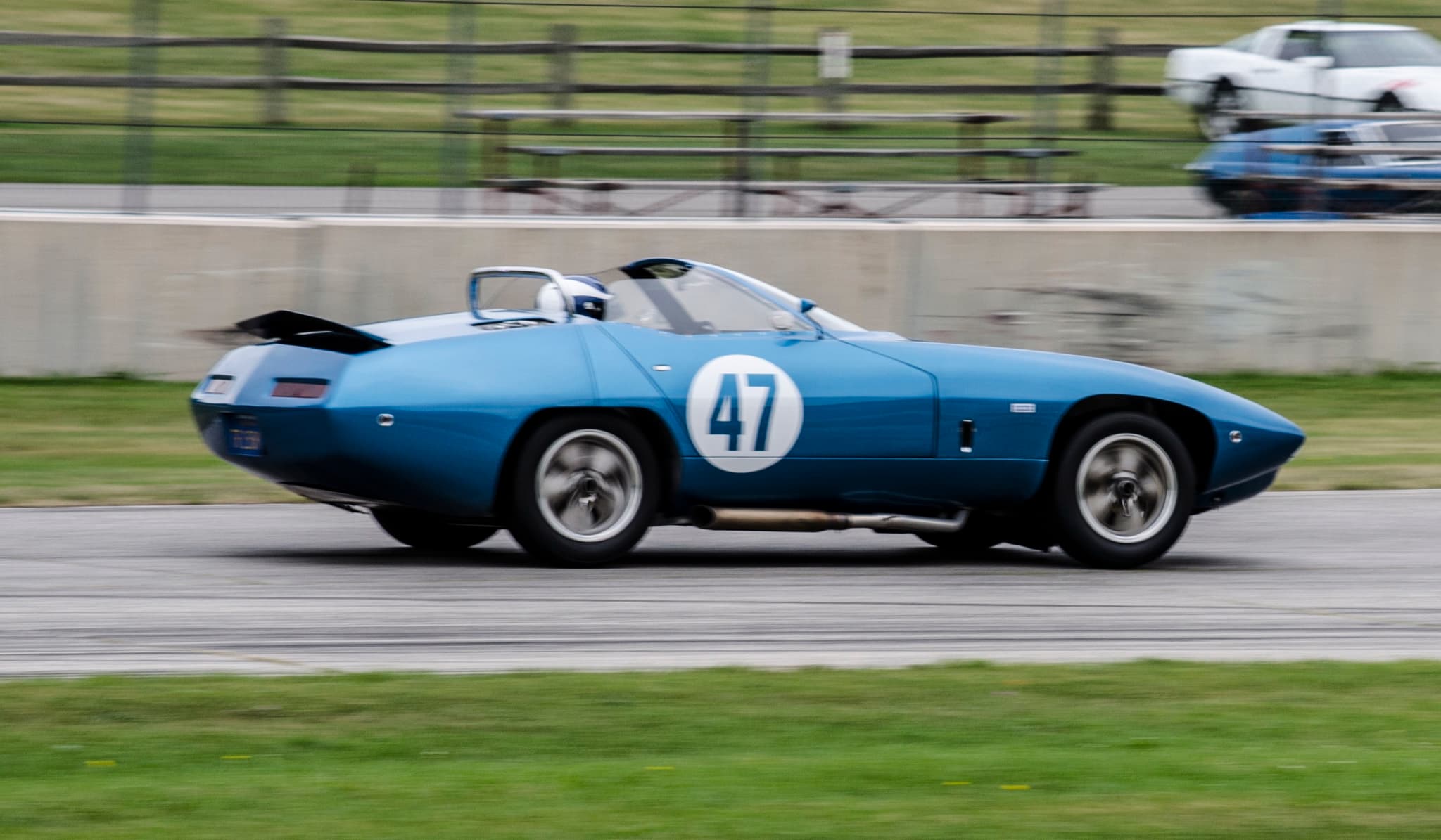 A low blue racing car with a long tail and number 47 in a white roundel, at speed on a circuit.