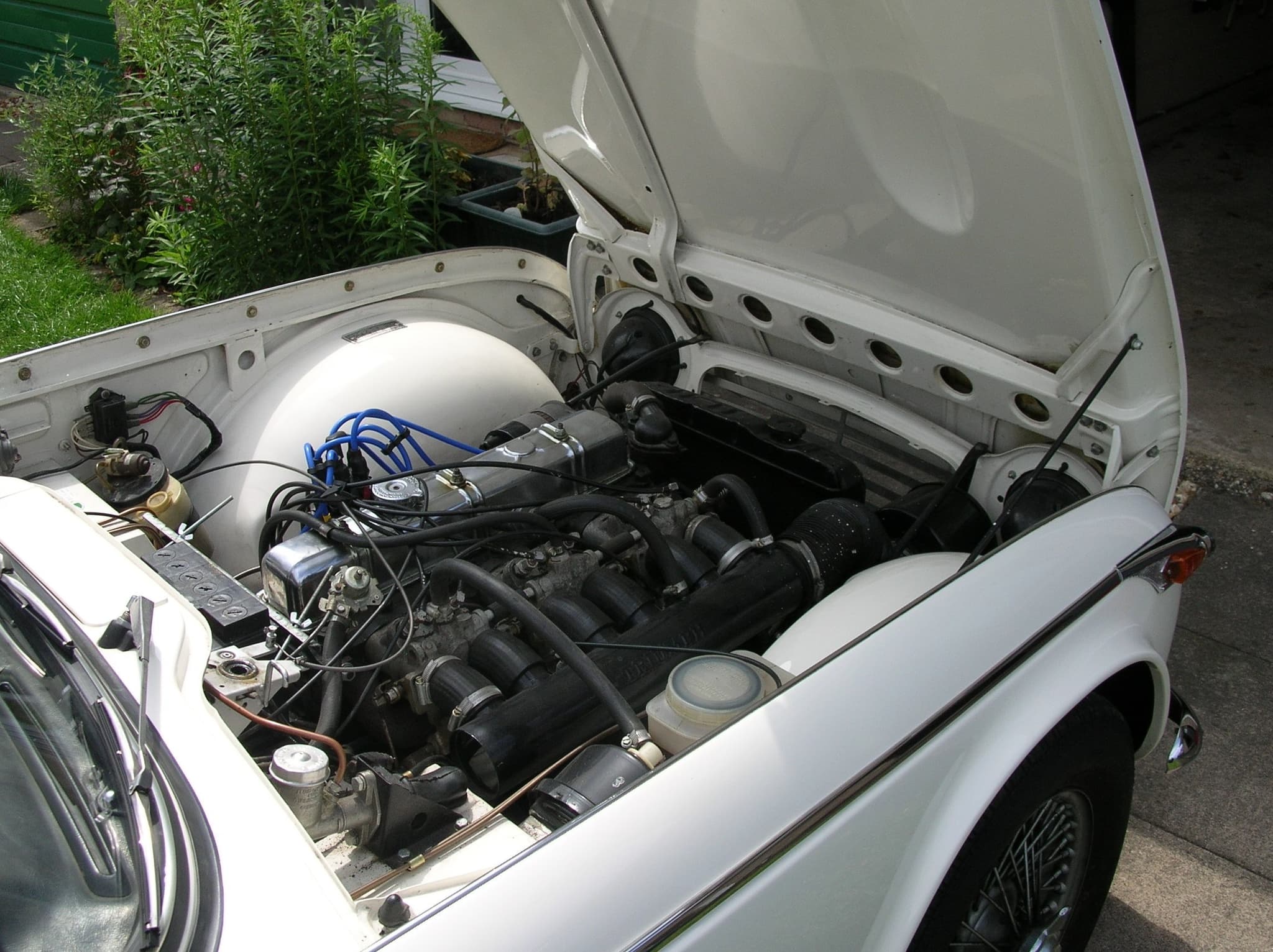 The engine bay of a white car with the bonnet raised: a six-cylinder engine with a long black inlet plenum and six short intake pipes.