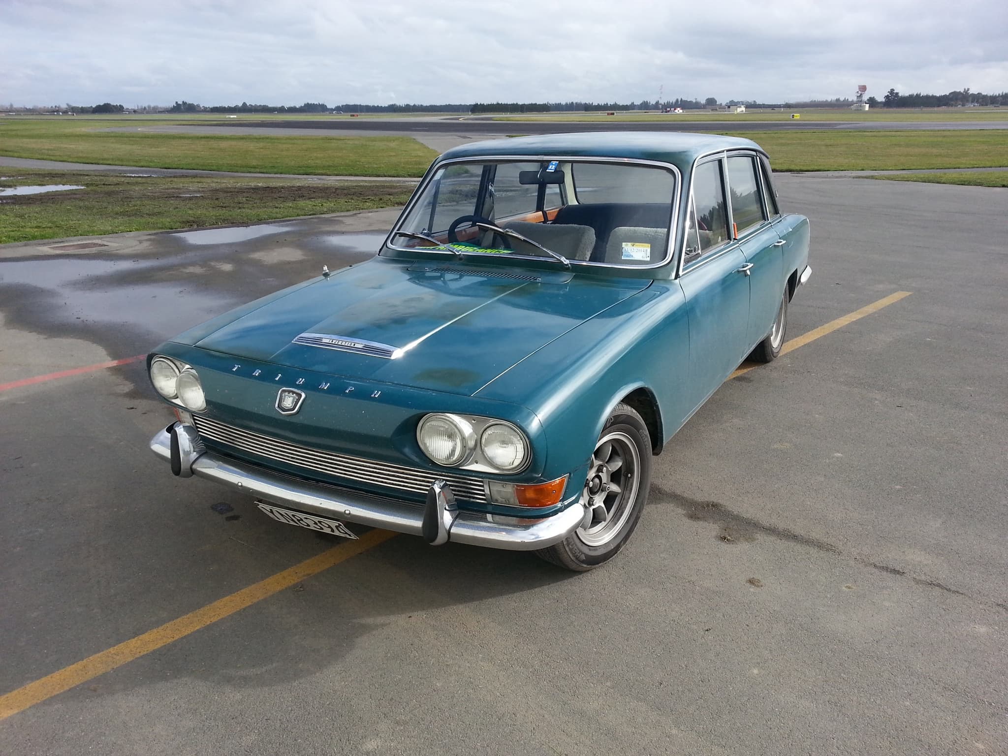 A blue four-door saloon with four headlamps, parked on a wet airfield apron under an overcast sky.