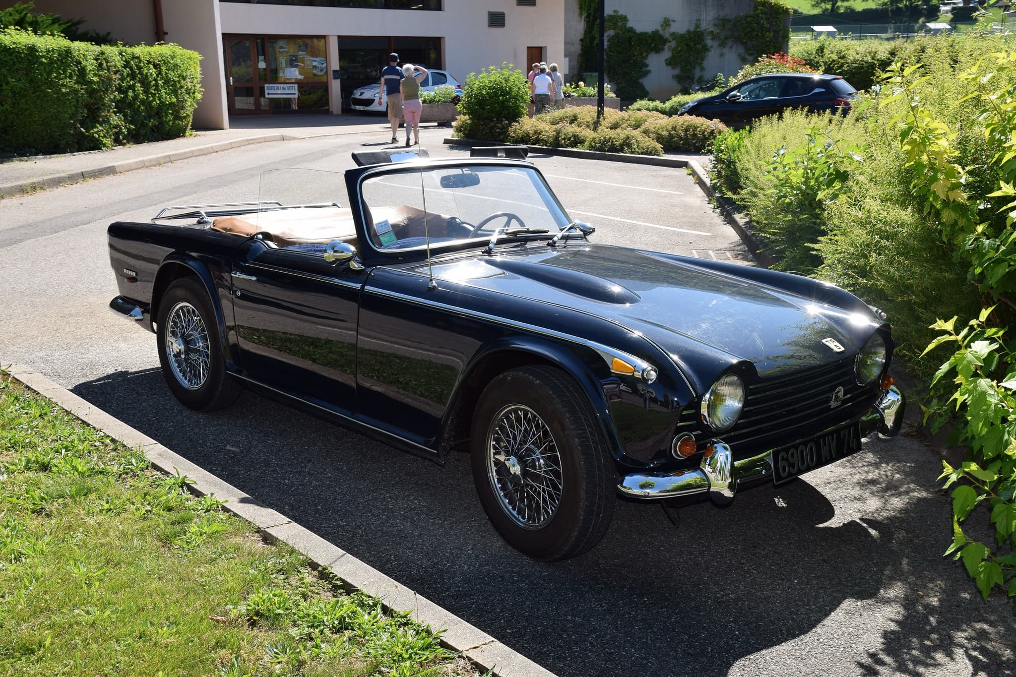 A dark blue sports car with wire wheels and its hood folded, parked in a sunny car park beside a hedge.