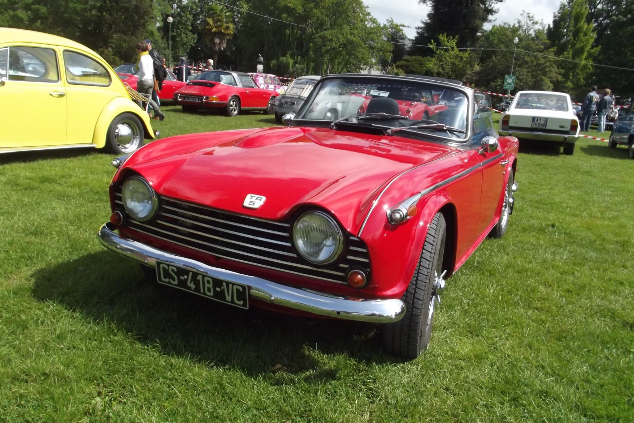 A red sports car with its black hood up on grass at a show, a yellow Beetle and other classic cars behind.