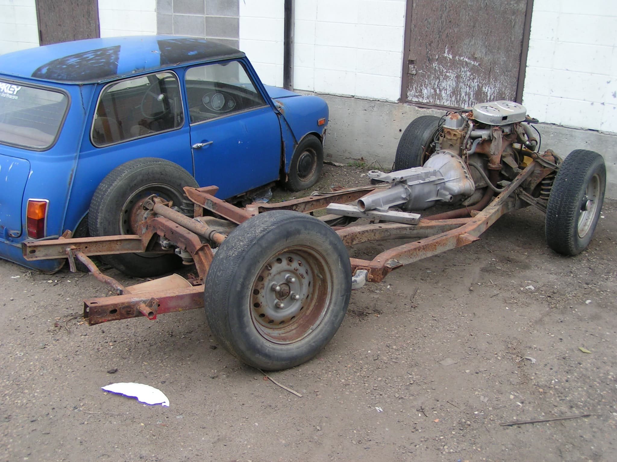 A rusty bare chassis frame on its wheels in a yard, with the six-cylinder engine, gearbox and rear suspension still fitted.