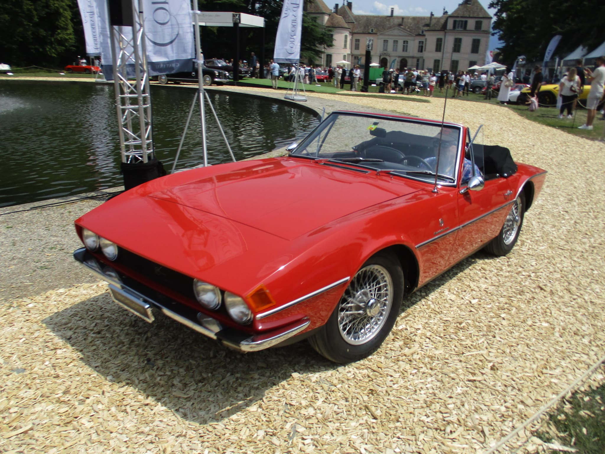 A red wedge-nosed roadster with four headlamps behind a clear cover and wire wheels, on gravel beside a pond at a concours.