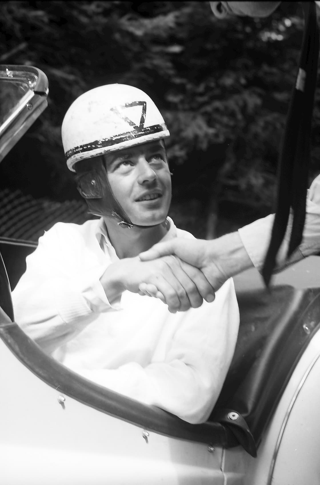 A racing driver in a white helmet numbered 7 sits in an open car and shakes an outstretched hand.