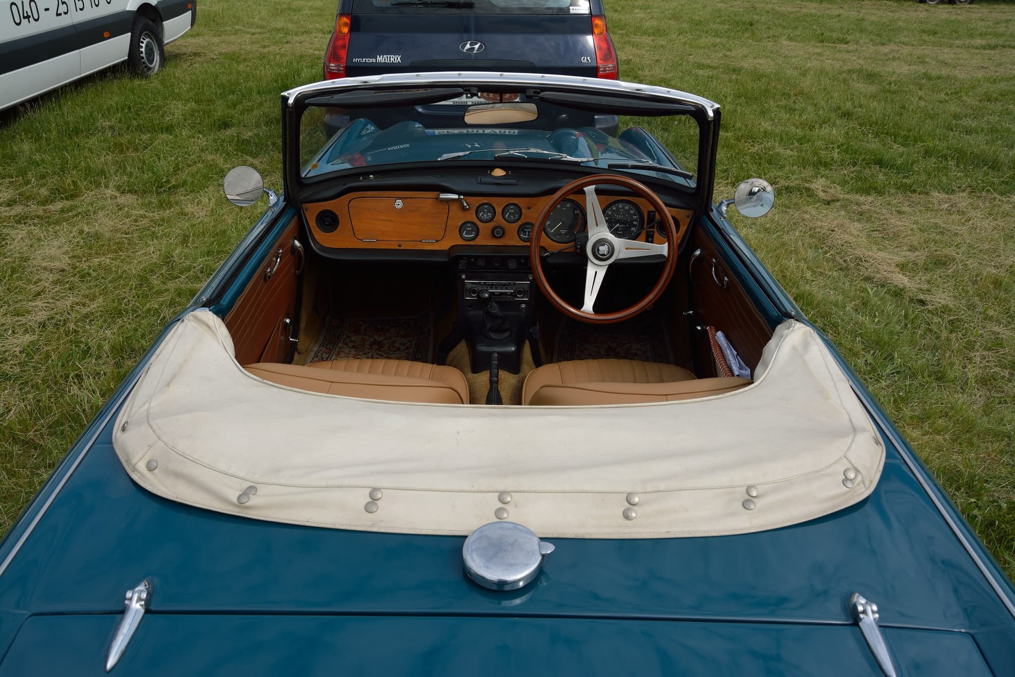The cockpit of a teal open car seen from behind: a wooden dashboard, a wood-rimmed wheel and tan seats behind a cream hood cover.